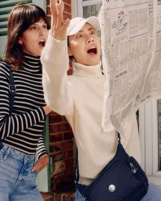 Two people in front of a house, one holding a newspaper and the other with a backpack.