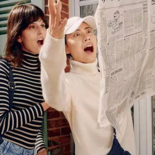 Two people in front of a house, one holding a newspaper and the other with a backpack.