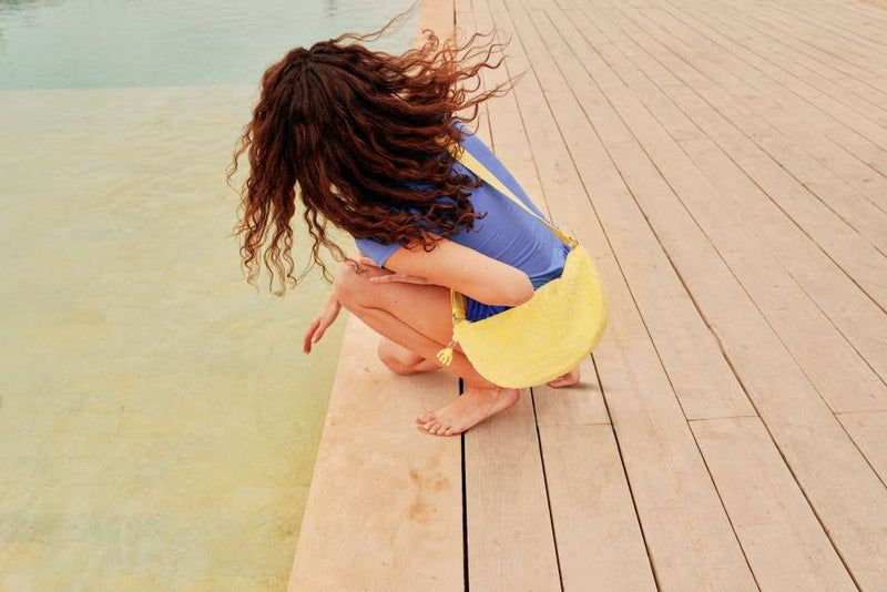 Woman in a blue swimsuit with a yellow bag sitting on a wooden dock by a body of water.