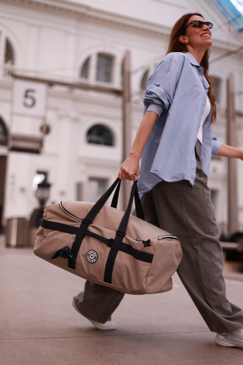 Woman walking outdoors holding a beige duffel bag with a building in the background